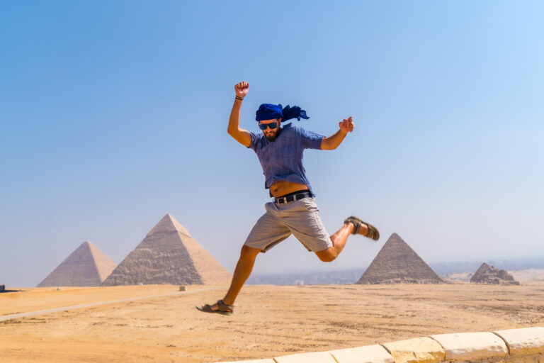 A young tourist jumping for joy in a blue turban and sunglasses at The Pyramids of Giza, the oldest Funerary monument in the world. In the city of Cairo, Egypt